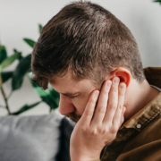 Unhealthy sad caucasian man 30s suffering from painful otitis. Health care, medicine and tinnitus concept. Closeup up of upset young beaded male having ear pain touching painful head sitting on sofa Les acouphènes : un trouble auditif à ne pas négliger